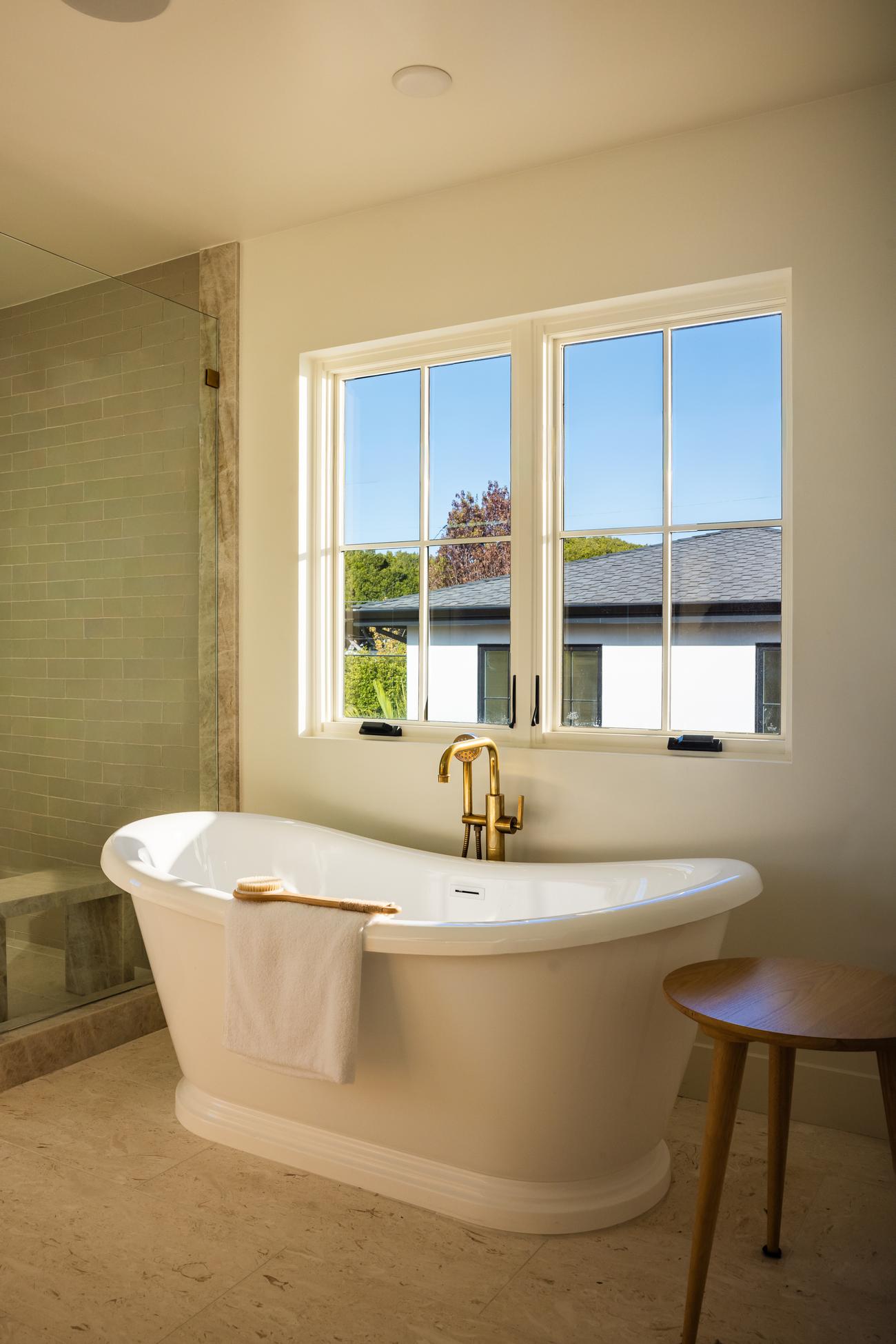 Freestanding soaking tub in the primary bath beneath a garden-view window, brass fixtures